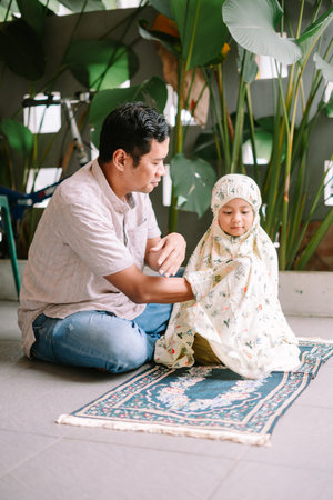 A Muslim father guides his young daughter during daily prayer, both sitting on a traditional prayer...