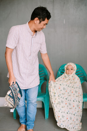 An adult Muslim man stands beside a young girl dressed in a floral prayer garment, holding a prayer...