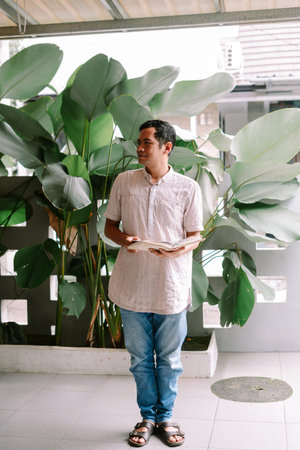 A man stands outdoors, holding an open book and looking away, with large green tropical plants and a...