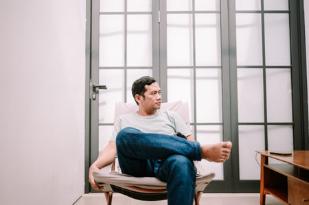 A thoughtful Asian man sits comfortably cross-legged in a modern chair indoors, looking away. He is...