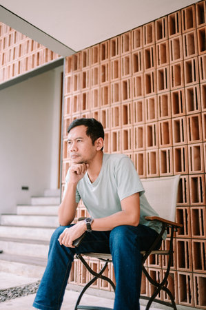 A casually dressed man sits thoughtfully, looking away, in a modern architectural setting featuring...