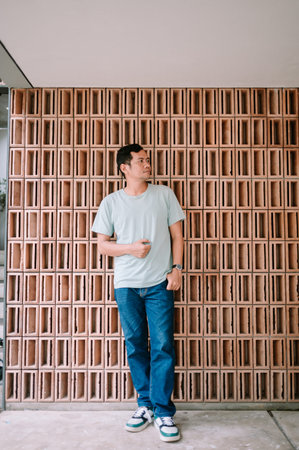 A man in a light t-shirt and jeans stands with a relaxed pose against a striking terracotta brick...