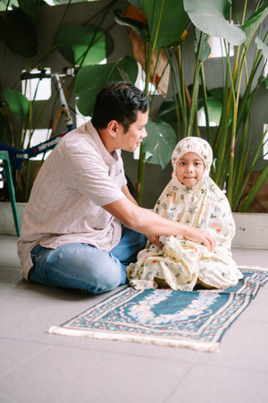 An adult Muslim man gently guides a young girl in a floral prayer outfit as she sits on a prayer mat, learning the ritual of prayer at home. This scene highlights intergenerational religiousの写真素材
