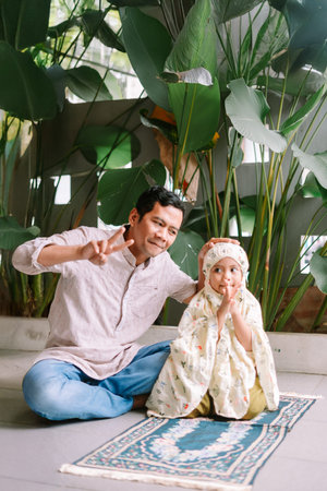 A father and his young daughter share a spiritual moment indoors. The girl wears a prayer garment and sits on a prayer mat, with her father nearby, emphasizing family bonds and religious practice.の写真素材