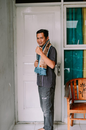 A friendly Asian man smiles at the camera, wearing a vibrant patterned scarf and casual clothing,...