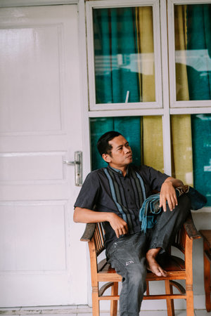 A contemplative Southeast Asian man sits casually in a traditional patterned shirt on a wooden...