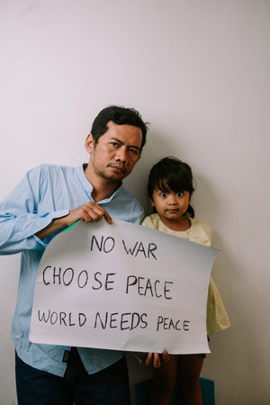 A father and his young daughter stand together, holding a powerful sign advocating for 'No War Choose Peace World Needs Peace'. Their serious expressions convey the urgency of this global message.の写真素材