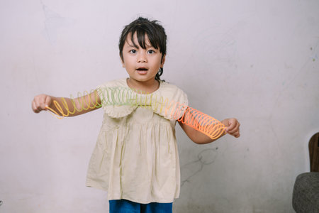 An adorable young girl expresses wonder while playfully stretching a classic rainbow spring toy,...