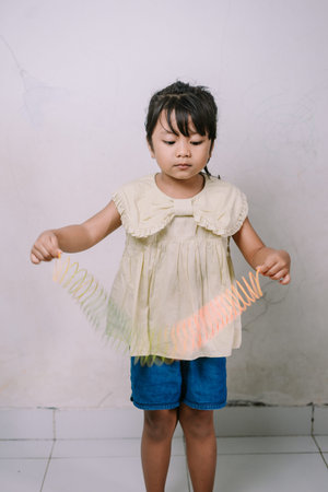 A young girl is happily playing with a vibrant, colorful coiled spring toy indoors. Her focused...
