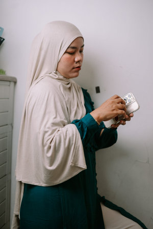 A young Muslim woman wearing a hijab is focused on her smartphone, browsing content or...