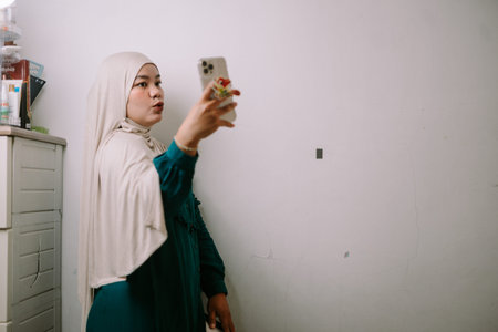 A young Muslim woman wearing a hijab and a green dress is actively recording herself with a...