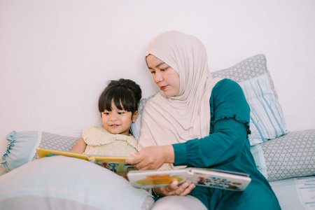 A caring Asian Muslim mother reading a children's book with her daughter, supporting early childhood...