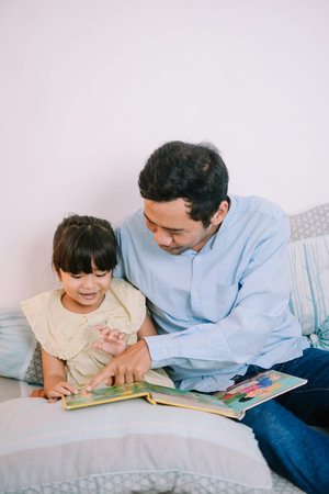 A young Muslim father helping his daughter understand a storybook, supporting early education and...