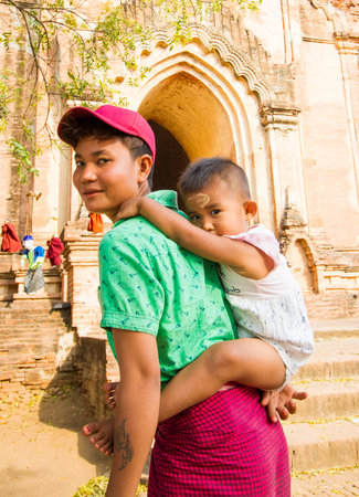 BAGAN,MYANMAR-MARCH 9:Burmese woman holding little girl on her back in front of ancient temple in Bagan,Myanmar on March 9,2017.Burmese children like to put Thanaka on their face.のeditorial素材