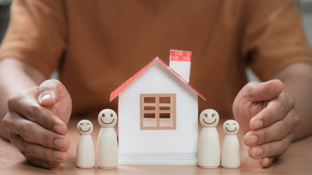 Concept of family protection and home security with hands shielding smiling wooden figures beside house model, symbolizing real estate stability and residential investment for future safety.の写真素材
