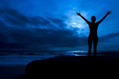 Midnight glory - female silhouette with arms raised, in a cloudy moonlit night, by the oceanの写真素材