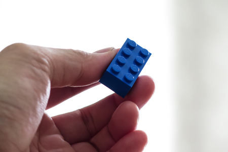 Hand holding a single blue Lego toy brick on a bright background.のeditorial素材
