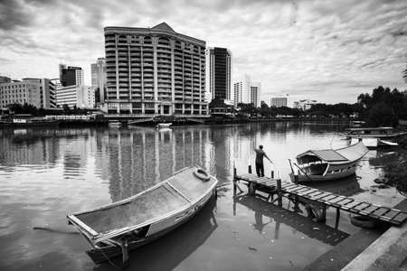 Black and white of a man waving hand with his boat to the other side of the river having buildings.のeditorial素材