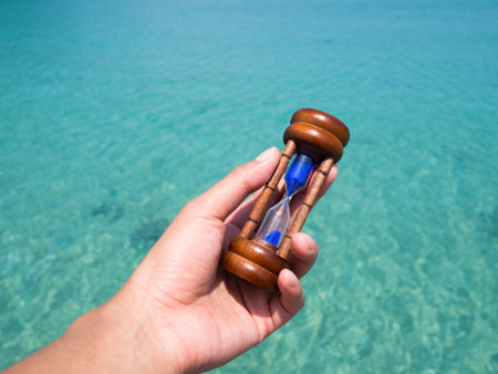 lady hand holding hourglass, sandglass against with green blue sea water surface in background. Time...