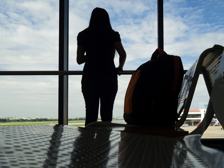 Silhouette woman stand and look outside window with bag on chair waiting flight in airport. Woman travel alone.の写真素材
