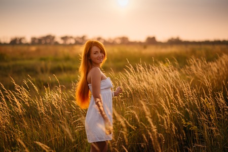 Young beautiful red-haired girl in a field at sunset, looking into the cameraの写真素材