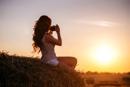 beautiful young redhead woman with vintage camera in a field at sunset, back to cameraの写真素材