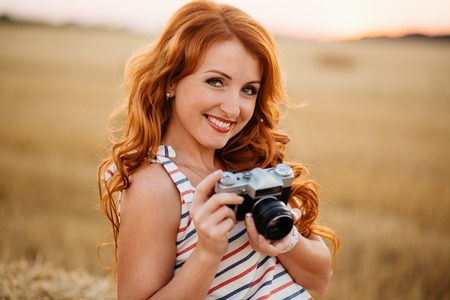 beautiful young redhead woman with vintage camera in a field at sunset, looking at camera, smilingの写真素材