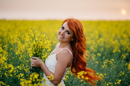 Young beautiful red-haired woman in a blooming rapeseed field at sunsetの写真素材