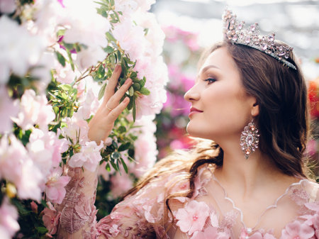 Beautiful young woman with a crown on her head and in a peach-colored dress walking in a blooming garden, close-upの写真素材
