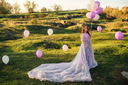 Red-haired woman with balloons in her hand and in a fairy purple dress is standing on the green lawnの写真素材