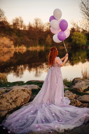 Red-haired woman with balloons in her hand and in a fairy purple dress is standing by the river at sunsetの写真素材