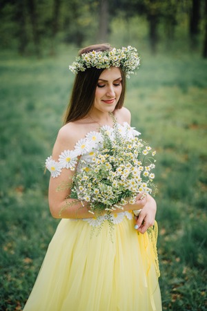 A young woman dressed in a beautiful tender dress and a wreath holds a bouquet of chamomiles in her handsの写真素材