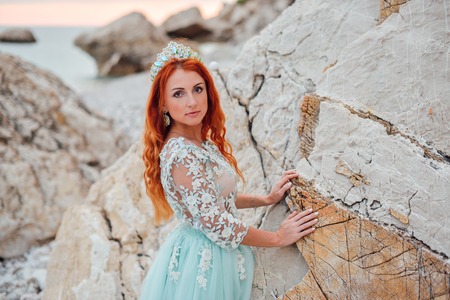 Young beautiful red-haired woman in a luxurious dress stands on a rocky shore of the Adriatic Sea among large stones, close-upの写真素材