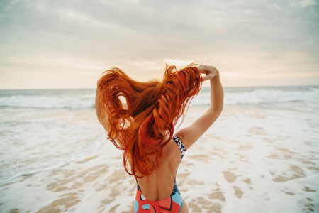 young red-haired woman with flying hair on the ocean coast at sunset, rear viewの写真素材