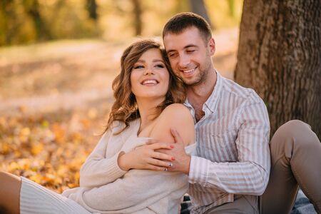 Smiling woman in the arms of her beloved is sitting in the autumn park by the tree on a sunny day.の写真素材