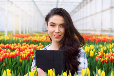 Young smiling female florist is standing with a tablet in her hands in a greenhouse. Close-up.