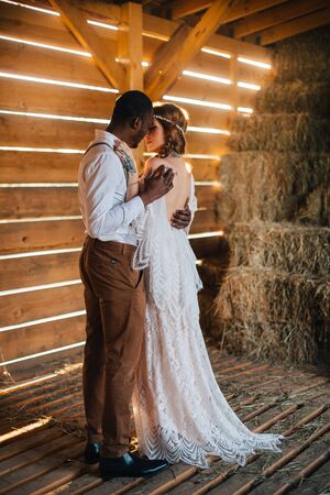 Newlyweds in the style of boho. African groom hugs a Caucasian bride in a barn in the sun. Full height portrait.の写真素材