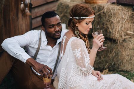 Boho style wedding. African-American bridegroom and Caucasian bride drink wine from glasses on a picnic.の写真素材