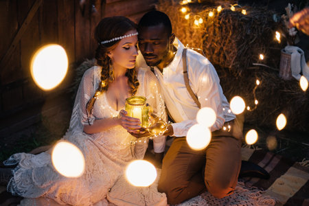 Boho style wedding. African groom and Caucasian bride sit embracing on a plaid surrounded by...