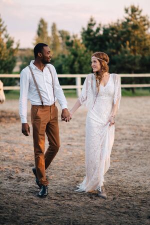 An interracial couple of newlyweds in the style of boho walk on a ranch at sunset. Soft focus.の写真素材