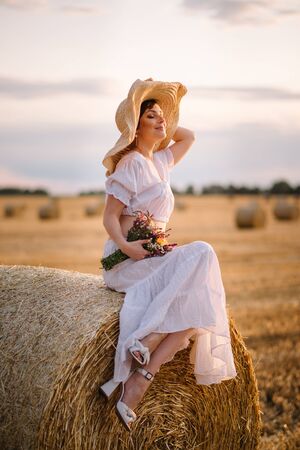 A young woman in a white sundress sits on a haystack on a mown field on a warm summer day.の写真素材