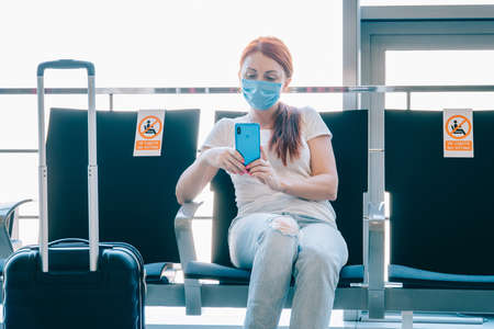 Social distance. Woman traveler uses a smartphone while waiting for a flight at the airport terminal. Stickers on adjacent seats with the inscription - no sitting.の写真素材