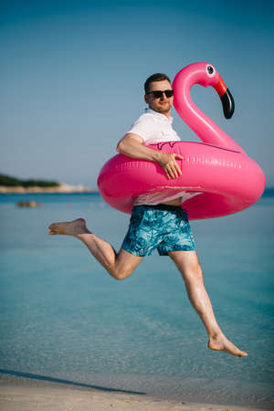 Summer vacation. Joyful male tourist in an inflatable pink flamingo is jumping up on the beach on a sunny day.の写真素材