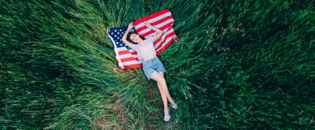 US Independence Day. Joyful teenager girl lies in the grass on the USA flag. Top down view.の写真素材