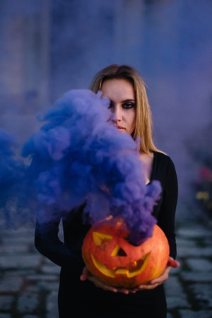 A young witch holds a steaming pumpkin with a terrible face in her hands. The smoke covers half of...