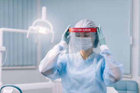 A young woman doctor dentist in a protective suit puts a protective shield on her face for additional protection during an epidemic.の写真素材