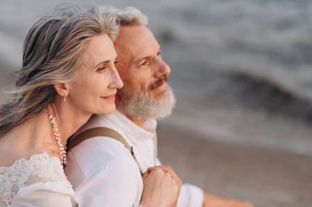 Romantic senior couple. An elderly couple in love is sitting on beach. Woman hugs man from behind. Close up.の写真素材
