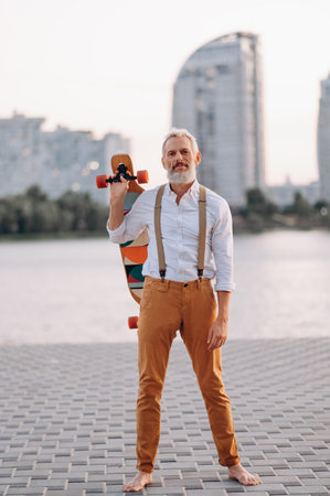 Gray-haired man holds skateboard in his hands and looks at camera. Full-length portrait.