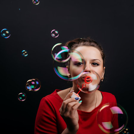 Young attractive woman in red dress blowing soap bubbles on black background.