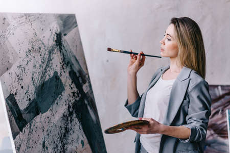 A young woman artist thoughtfully paints an oil painting in the style of abstract art in the workshop.の写真素材